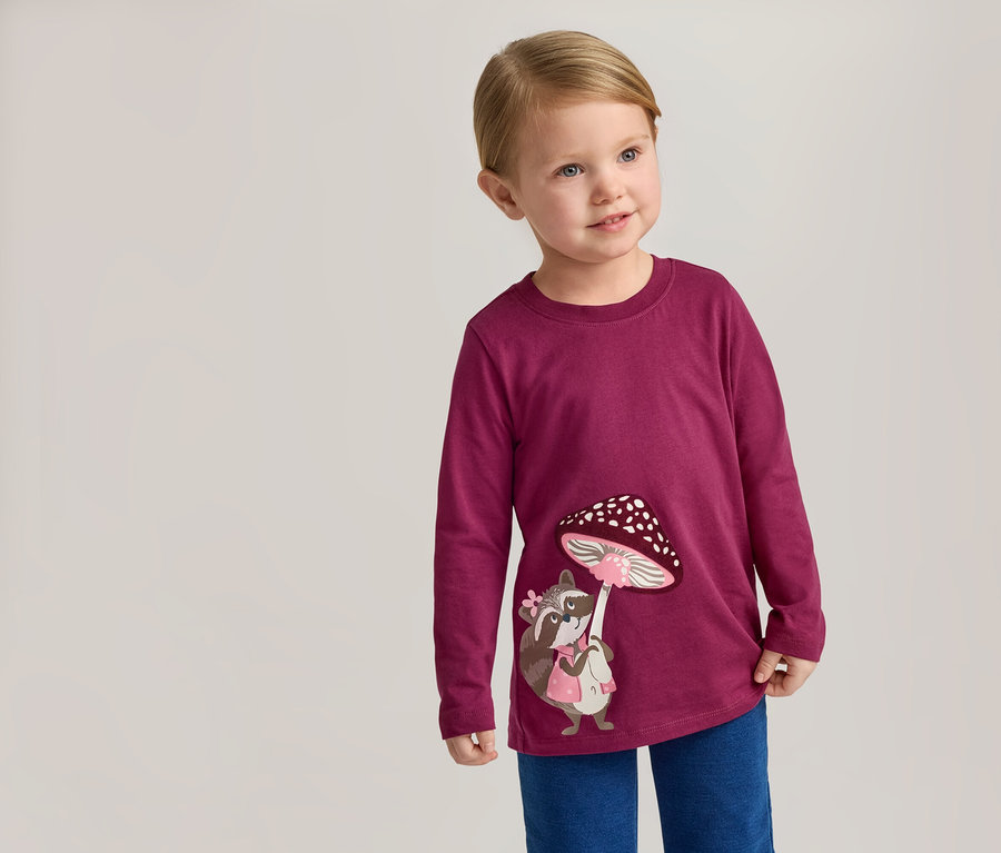 Ein blondes Kind trägt ein weinrotes Langarmshirt mit einem Waschbär und einem Pilz darauf und blaue Hosen.