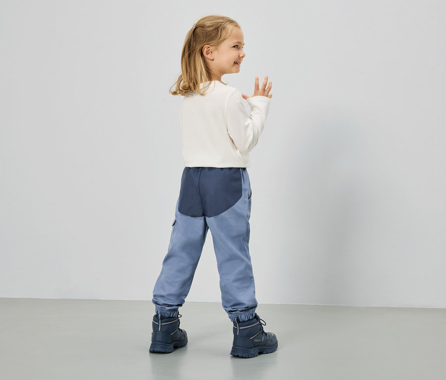 Ein kleines Mädchen mit blonden Haaren, gekleidet in ein weißes Langarmshirt, blaue Hosen und marineblaue Stiefel, steht vor einer weißen Wand.