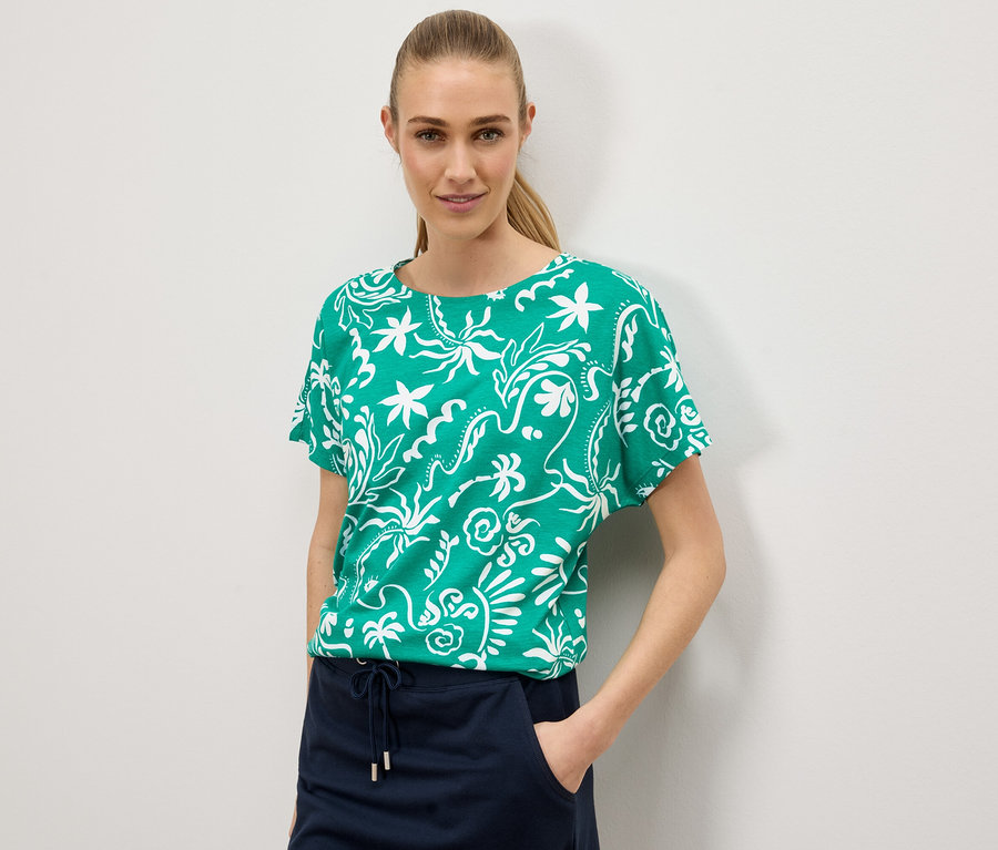 Frau trägt ein grünes T-Shirt mit weißem Blumenmuster und einen marineblauen Rock vor einem weißen Hintergrund.