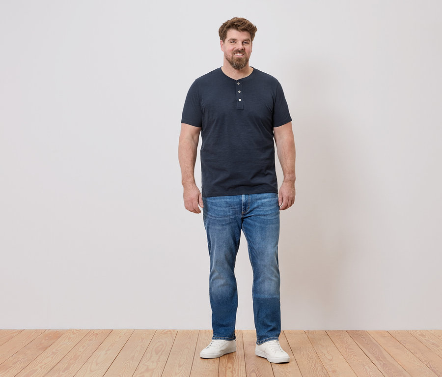 Ein Mann steht vor einer Wand, er trägt ein blaues T-Shirt, Jeans und weiße Schuhe.