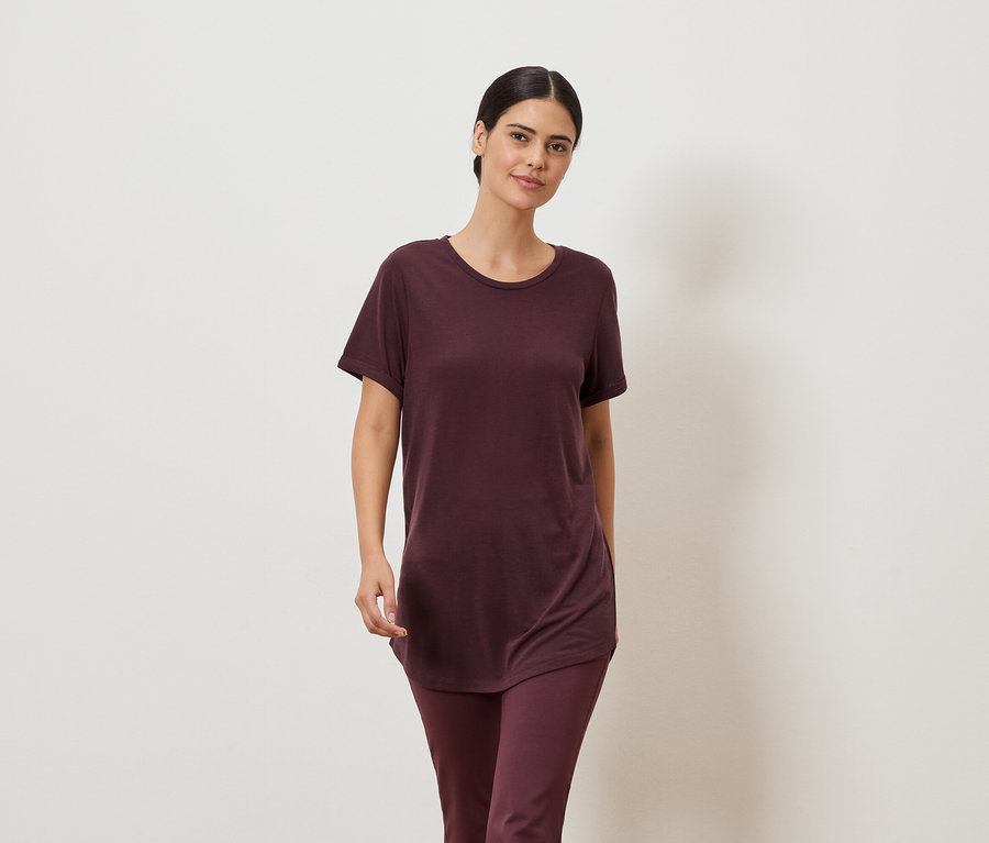 Frau in weinrotem T-Shirt und Leggings vor einem neutralen Hintergrund.