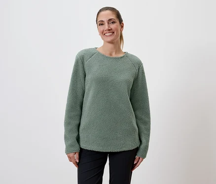 Eine lächelnde Frau mit Pferdeschwanz trägt einen grünen Fleecepullover und schwarze Hosen vor einem weißen Hintergrund.