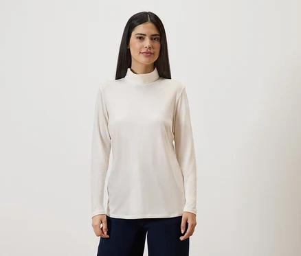 Frau mit langen, dunklen Haaren trägt ein langärmliges, cremefarbenes Rollkragenshirt und eine dunkle Hose vor einem hellen Hintergrund.