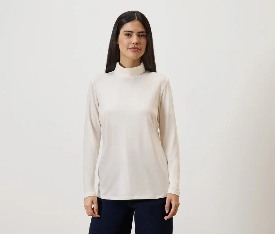 Frau mit langen, dunklen Haaren trägt ein langärmliges, cremefarbenes Rollkragenshirt und eine dunkle Hose vor einem hellen Hintergrund.