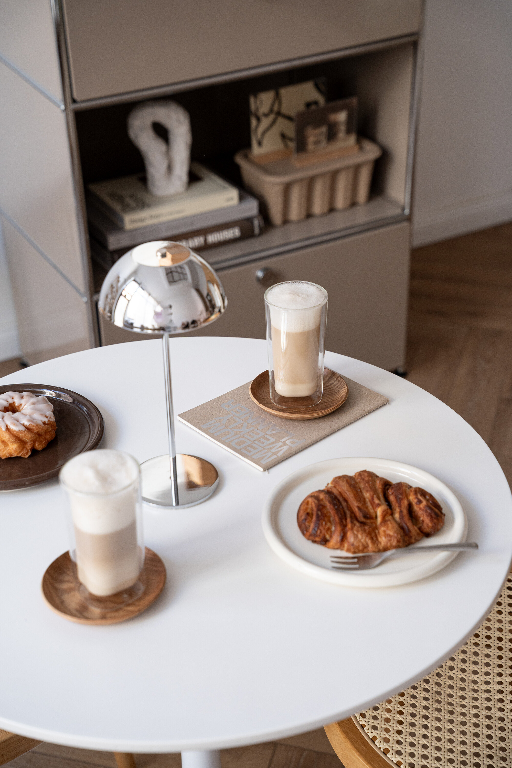 Auf einem Tisch stehen zwei Latte Macchiato, ein Croissant, ein Krapfen und eine Lampe.