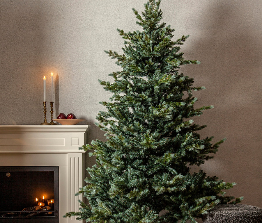 Ein ungeschmückter Weihnachtsbaum steht auf einem Holzboden, umgeben von roten Geschenken, einem grauen Schaffellteppich und einem dunkelgrauen Pouf. Links daneben befindet sich ein weißer Kamin mit leuchtenden Holzscheiten, auf dessen Sims zwei brennende Kerzen und eine Schale mit roten Äpfeln stehen.