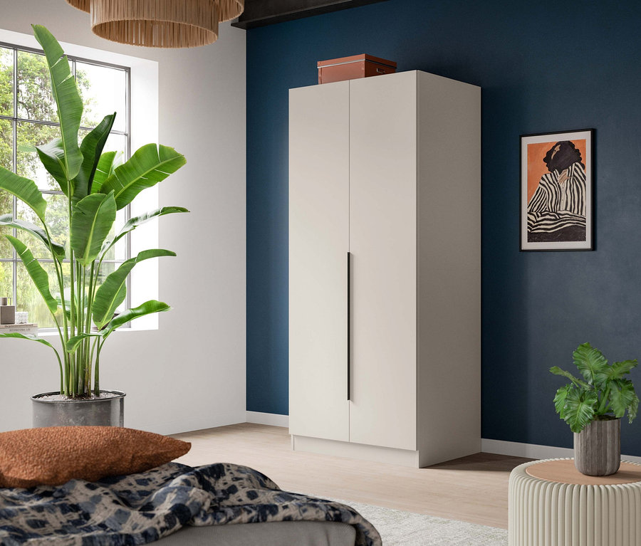 Modernes Schlafzimmer mit hohem beigem Kleiderschrank vor dunkelblauer Wand. Eine große Grünpflanze steht am Fenster, ein Bett mit gemusterter Decke ist im Vordergrund. Ein gerahmter Kunstdruck hängt an der Wand, eine kleine Pflanze steht auf einem gerippten Hocker.