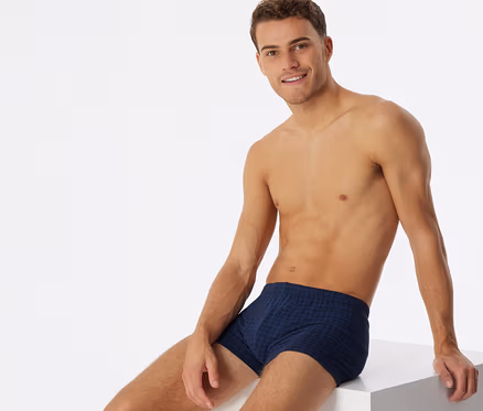 Oberkörperfreier Mann in dunkelblauen, karierten SCHIESSER Shorts, lächelnd auf einem weißen Würfel sitzend.