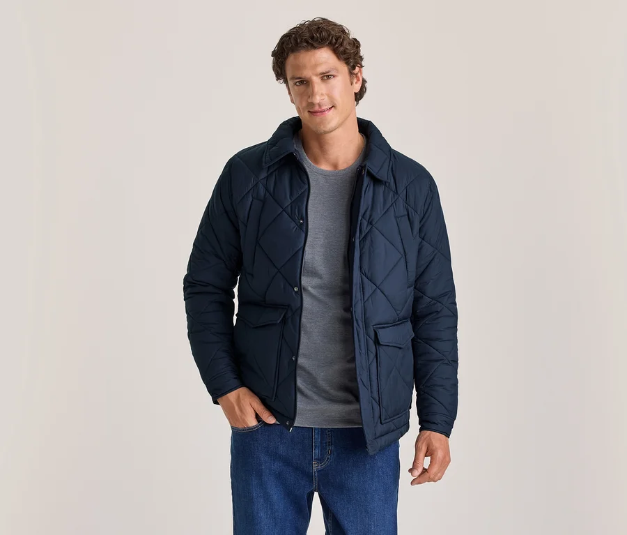 Mann in einer dunkelblauen Steppjacke über einem grauen T-Shirt und blauen Jeans, der mit einer Hand in der Tasche steht und in die Kamera blickt.