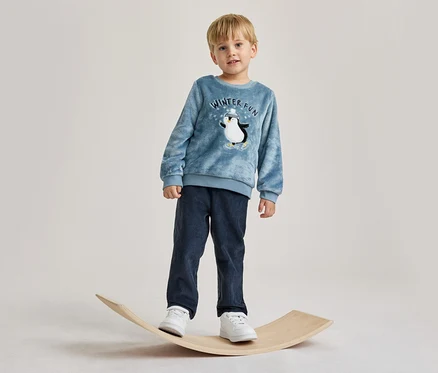 Junger Junge steht auf einem hölzernen Balance Board. Er trägt ein blaues Langarmshirt mit Pinguin-Motiv und „WINTER FUN“-Schriftzug, eine dunkelblaue Cordhose und weiße Turnschuhe.