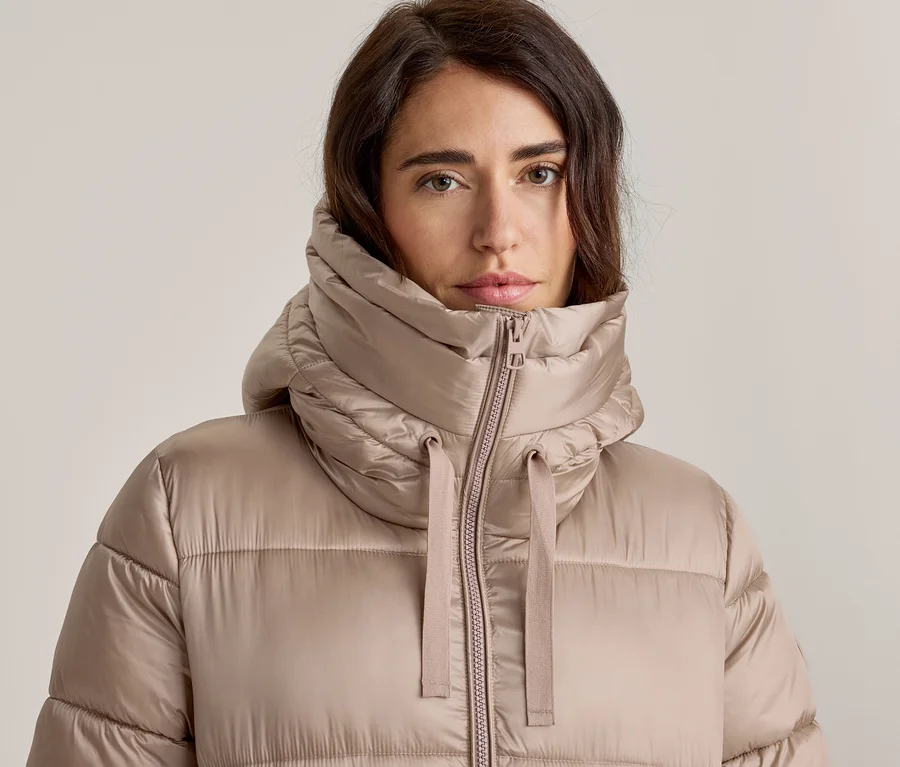 Nahaufnahme einer Frau mit dunklem Haar, die eine hellbraune Steppjacke trägt. Die Jacke hat einen hohen, gepolsterten Kragen mit Reißverschluss und sichtbaren Zugbändern, und sie blickt direkt in die Kamera.