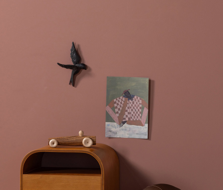 Eine altrosafarbene Wand mit einer schwarzen Vogeldekoration und einem abstrakten Gemälde. Darunter ein heller Holznachttisch mit einem Spielzeugauto und ein dunkelbrauner, kugelförmiger Pouf auf hellem Holzboden.