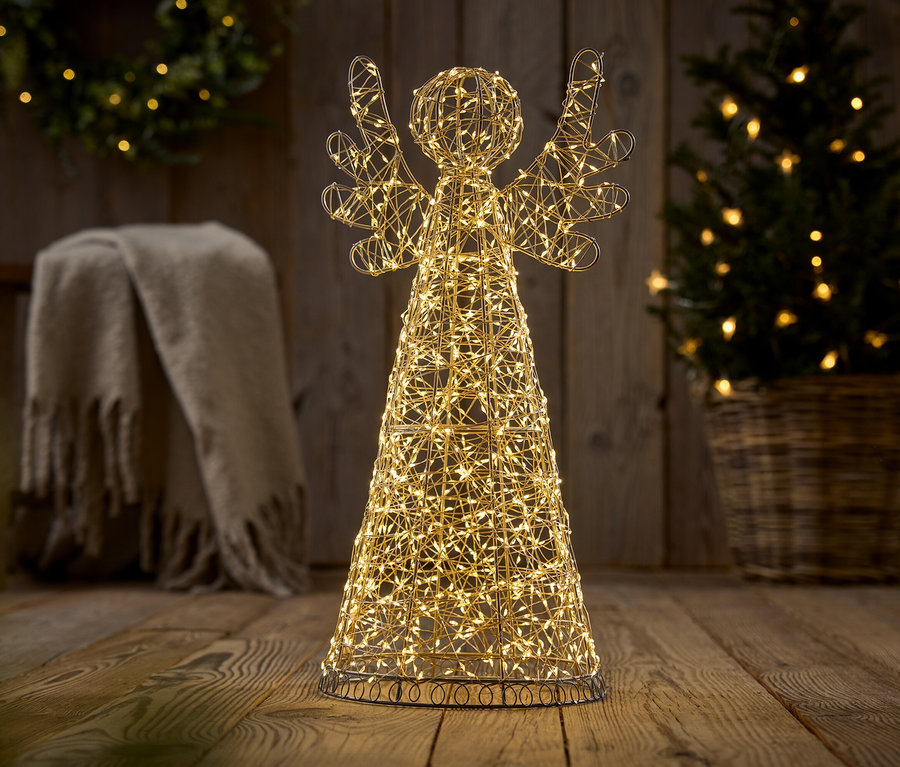 Leuchtende Draht-Engelsfigur mit warmweißen Lichtern auf Holzboden. Im Hintergrund ein beleuchteter Weihnachtsbaum in einem Korb und eine Decke auf einem Hocker.