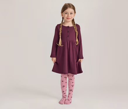 Ein junges Mädchen mit blonden Zöpfen lächelt stehend in einem langärmeligen pflaumenfarbenen Kleid mit Knöpfen und Taschen sowie rosa Strumpfhosen mit dunklem Blumenmuster.