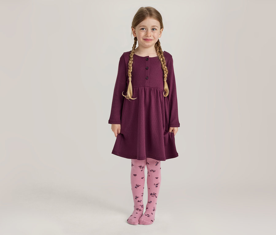 Ein junges Mädchen mit blonden Zöpfen lächelt stehend in einem langärmeligen pflaumenfarbenen Kleid mit Knöpfen und Taschen sowie rosa Strumpfhosen mit dunklem Blumenmuster.