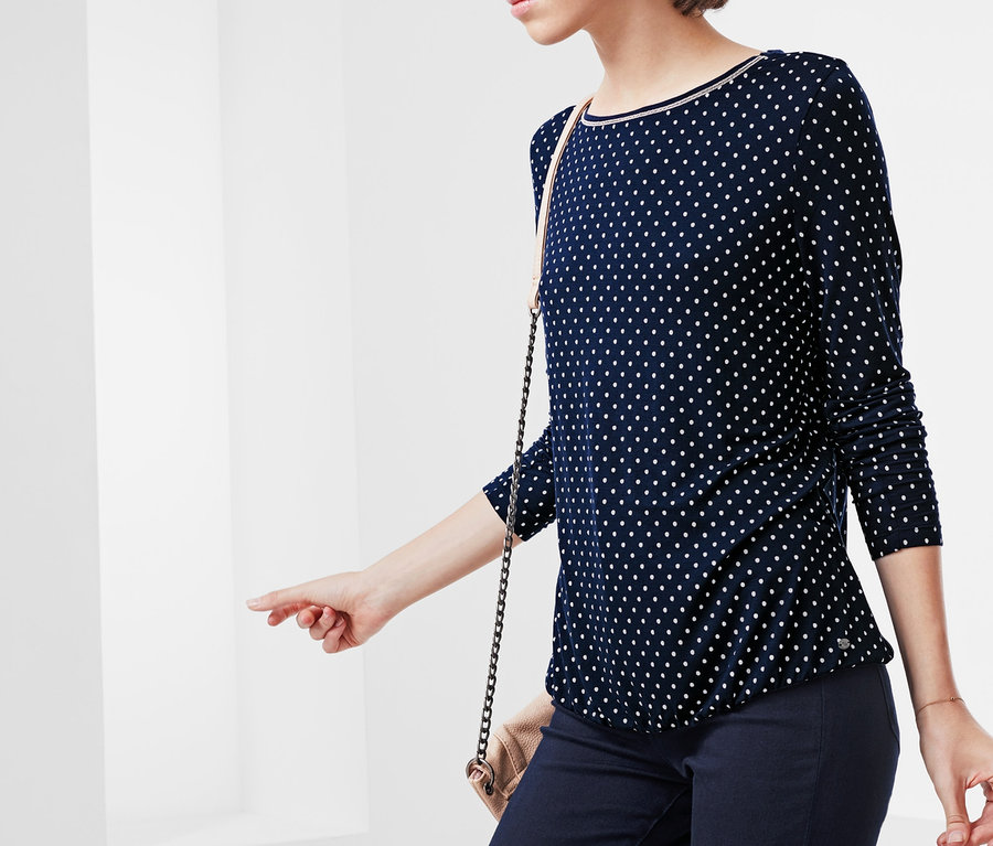 Frau mit lockigem Haar trägt eine blaue Bluse mit weißen Punkten und dunkelblaue Hosen.