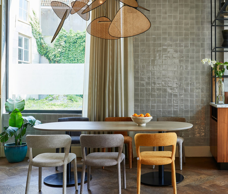 Ein modernes Esszimmer mit einem ovalen Holztisch, umgeben von sechs Stühlen in Hellgrau, Dunkelgrau und Senfgelb. Über dem Tisch hängt eine einzigartige Rattan-Deckenleuchte. Ein großes Fenster links blickt auf eine grüne Außenanlage, daneben steht eine Topfpflanze. Die rechte Wand ist mit hellgrauen Fliesen verkleidet, daneben ein dunkles Regal und ein Holzschrank mit weißen Blumen.