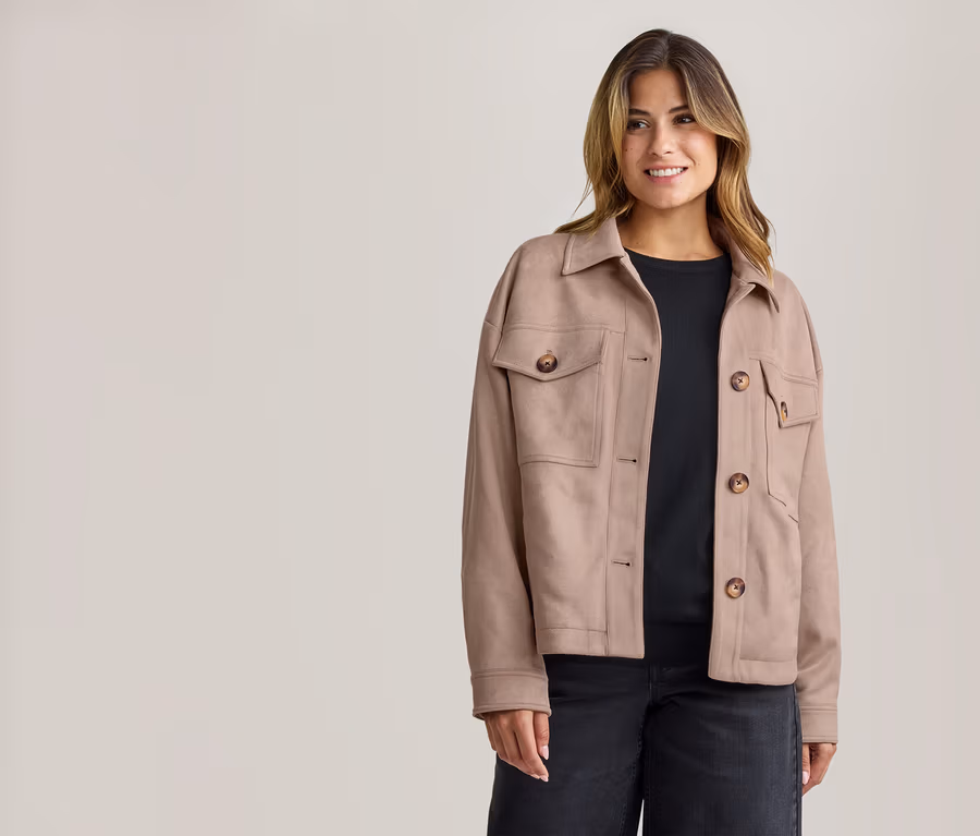 Lächelnde Frau in beiger Kragenjacke mit braunen Knöpfen und Taschen, schwarzem Oberteil und dunkler Jeans.