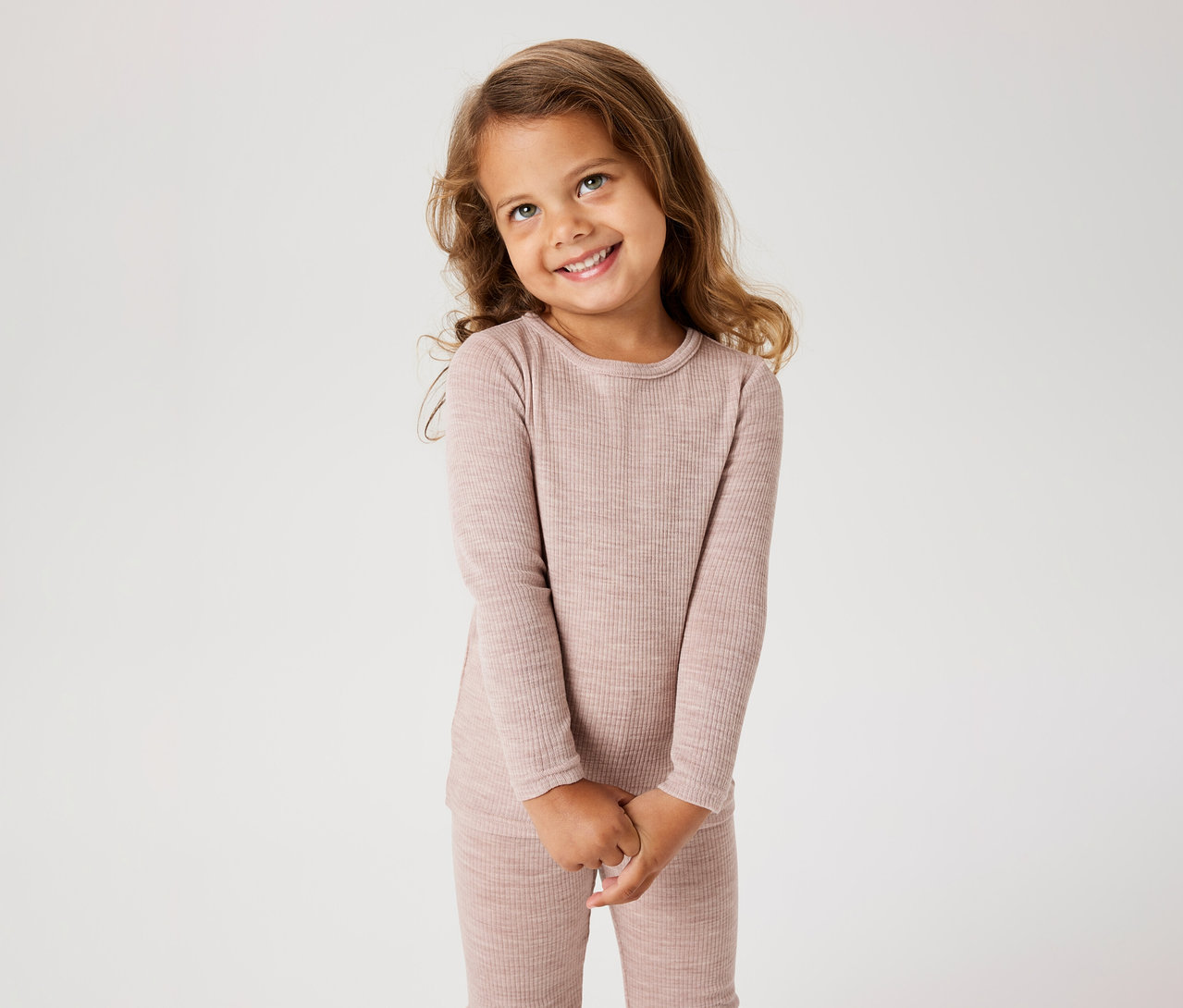Lächelndes kleines Mädchen mit langen braunen Haaren, das ein geripptes, hellrosa Langarmshirt und passende Hose trägt, die Hände gefaltet.