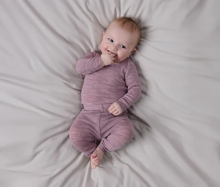 Lächelndes Baby liegt auf hellgrauem Laken, trägt langärmeliges, strukturiertes lila Oberteil und passende Hose, eine Hand am Mund.