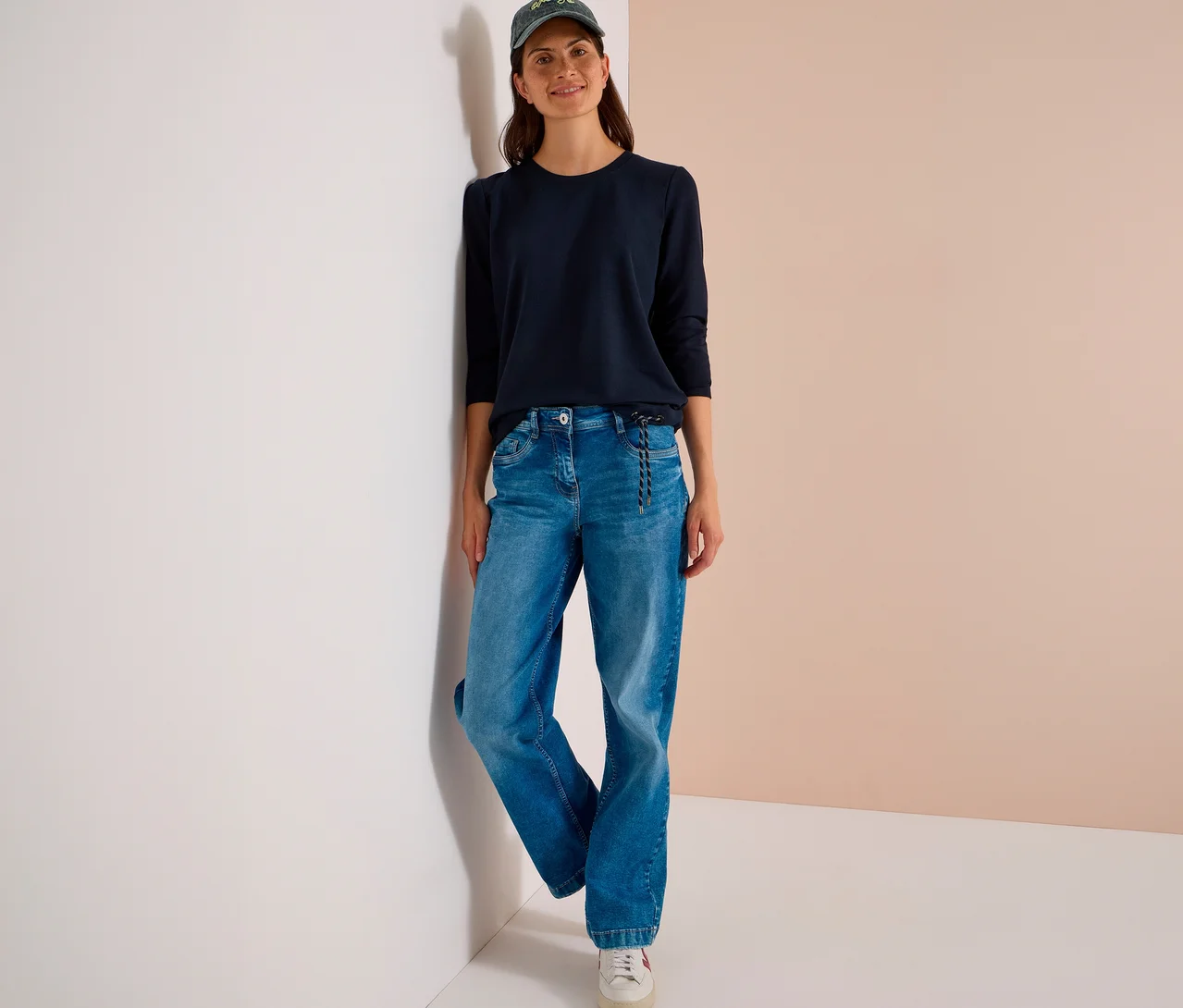 Eine Frau lehnt an einer Wand und trägt ein dunkelblaues CECIL Shirt, blaue Jeans, weiße Sneaker und eine grüne Baseballkappe.
