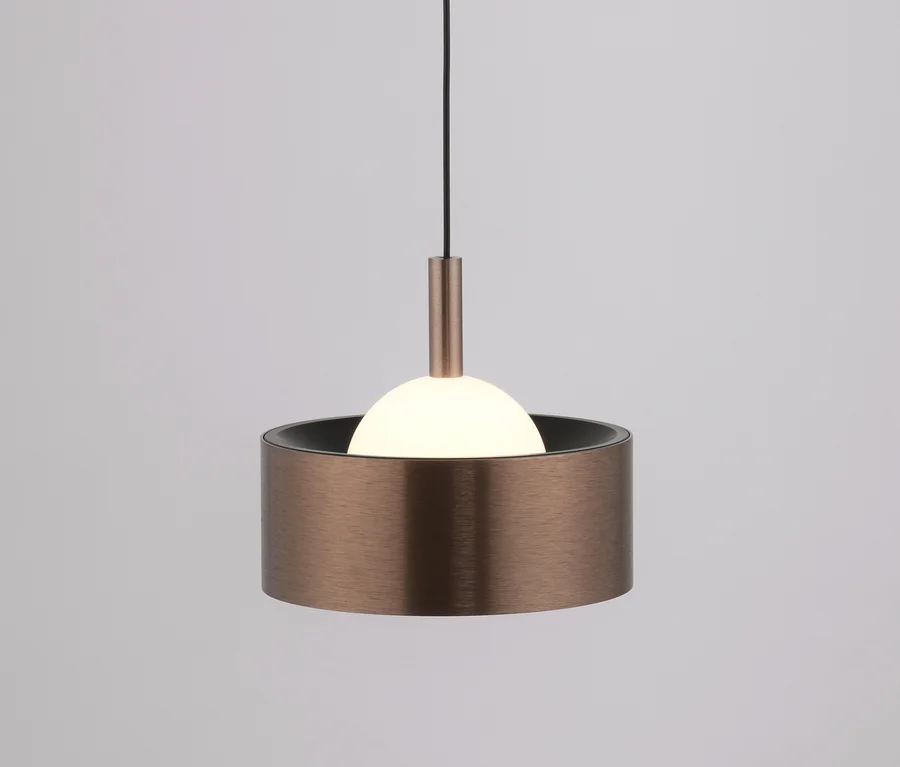 Eine moderne Pendelleuchte mit einem zylindrischen Schirm aus gebürsteter Bronze und einer leuchtenden weißen Kugelbirne, die teilweise sichtbar ist, an einem schwarzen Kabel vor hellgrauem Hintergrund.