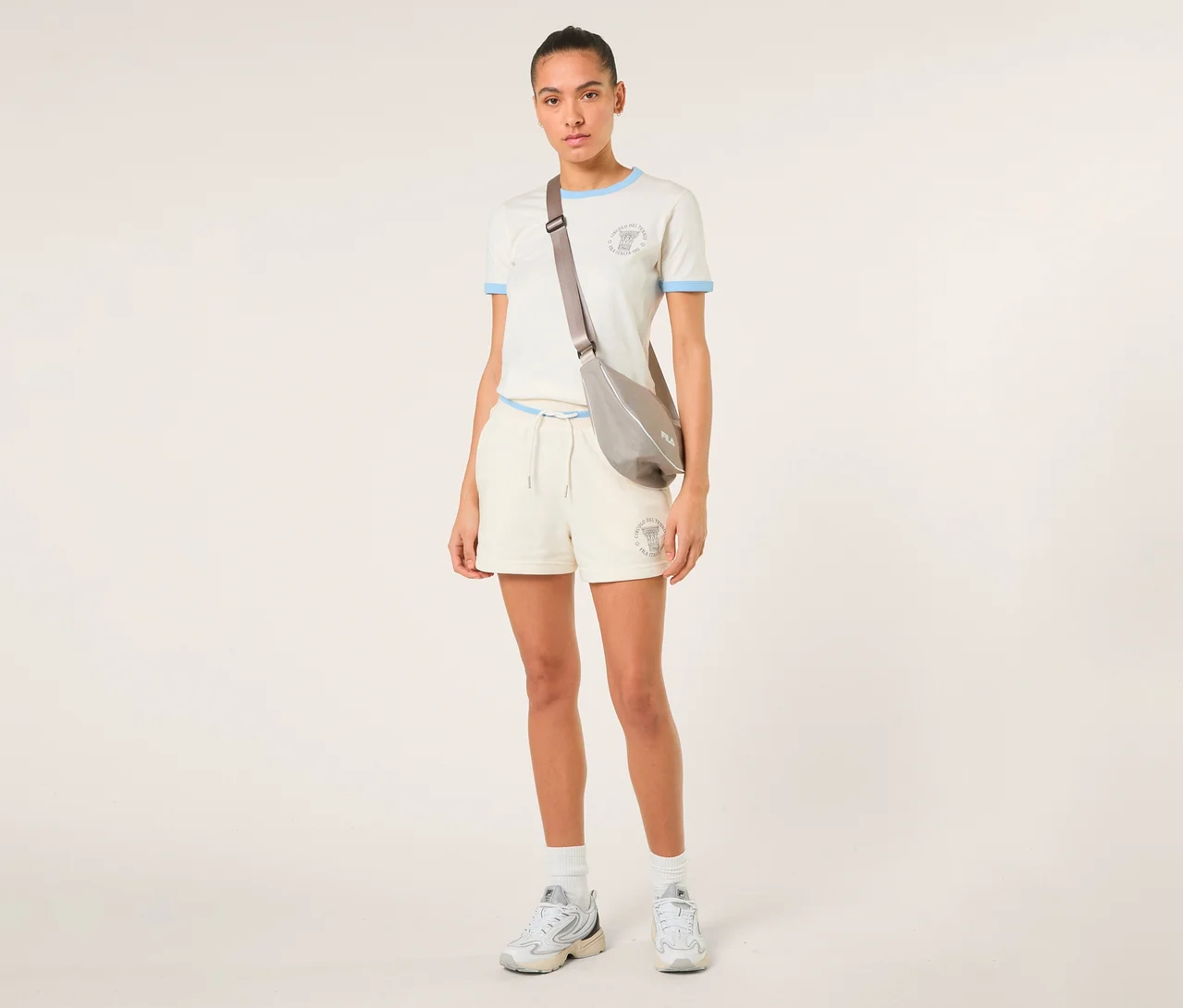 Eine Frau trägt offwhite FILA Damen Sweatshorts MARLIA, ein weißes T-Shirt mit hellblauen Akzenten, eine graue Umhängetasche und weiße Sneaker.
