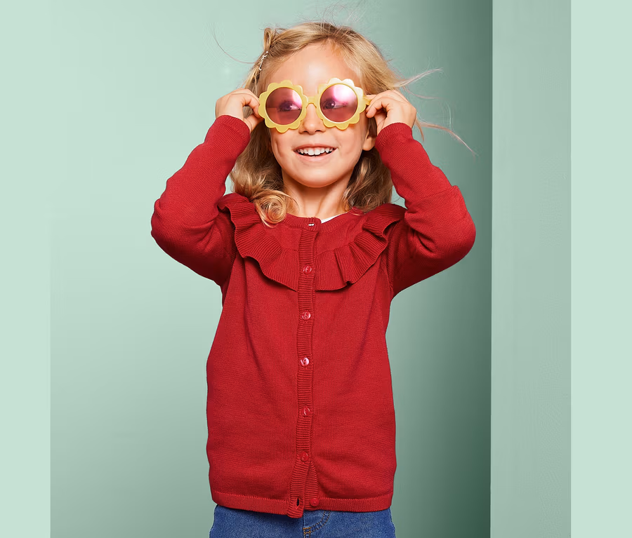 Mädchen mit blonden Haaren und gelber Sonnenbrille in Blumenform trägt eine rote Strickjacke und Jeans.