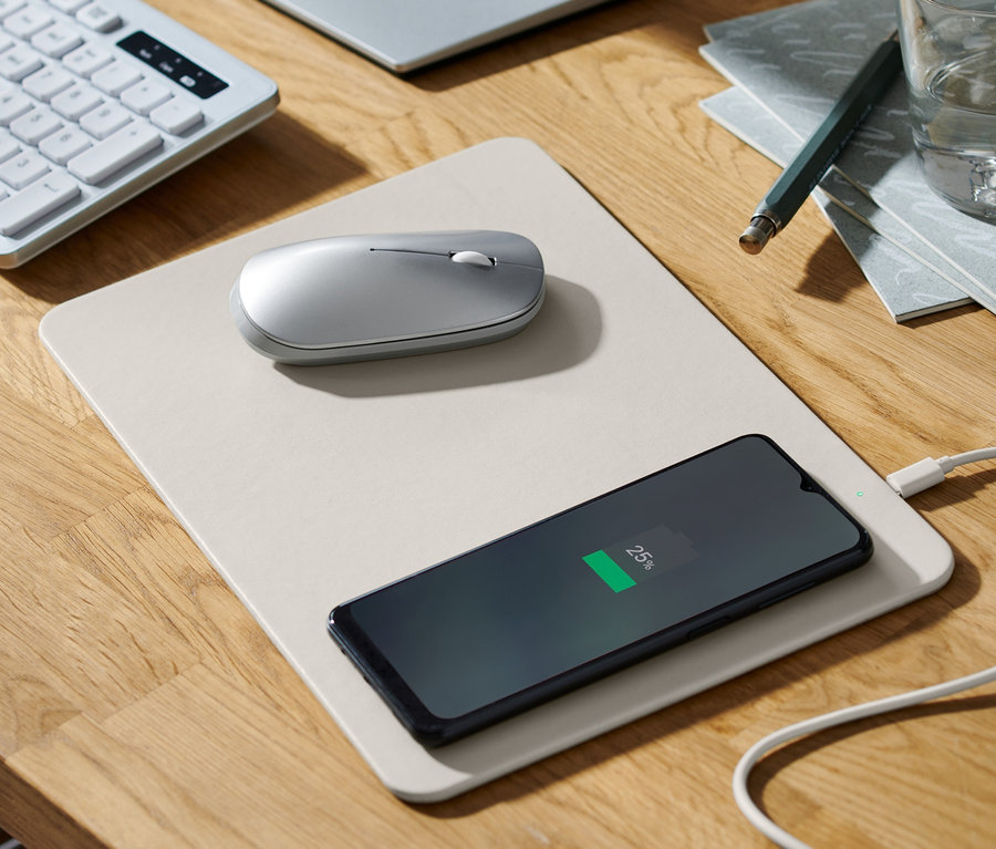 Auf einem Holztisch liegen ein Mauspad mit integriertem Ladegerät, auf dem ein Handy geladen wird, eine Maus, eine Tastatur, ein Monitor, ein Glas Wasser und ein Notizblock.
