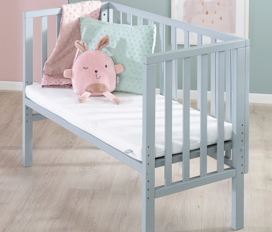 Graues Kinderbett mit rosafarbenem Plüschhasen, rosa Decke und grünem Kissen.