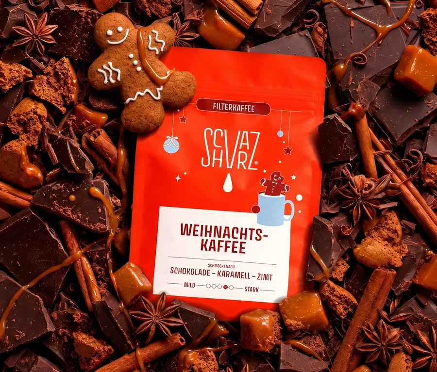 Eine rote Packung Schvarz Weihnachtskaffee Filterkaffee liegt inmitten einer festlichen Anordnung aus dunklen Schokoladenstücken, Karamellbonbons, Zimtstangen, Sternanis, Lebkuchenkrümeln und einem Lebkuchenmann, alles mit Karamellsauce beträufelt.