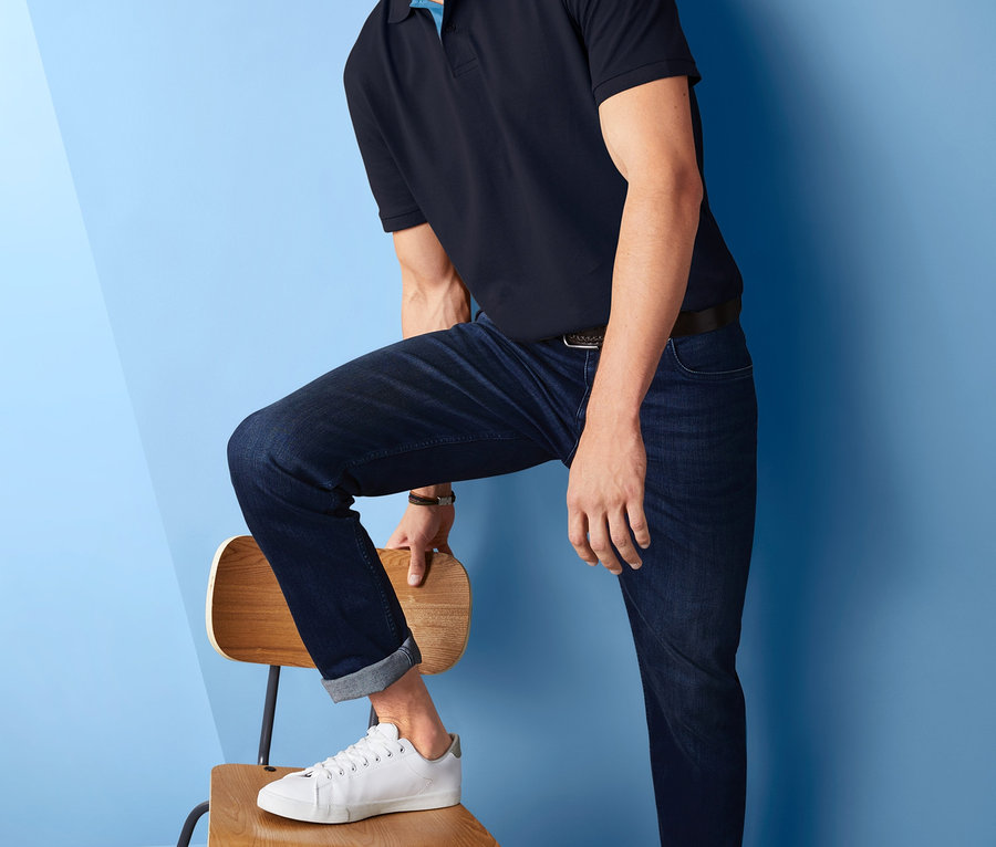 Lächelnder Mann posiert mit einem Bein auf einem Stuhl, er trägt ein marineblaues Piqué-Poloshirt, Jeans und weiße Turnschuhe.