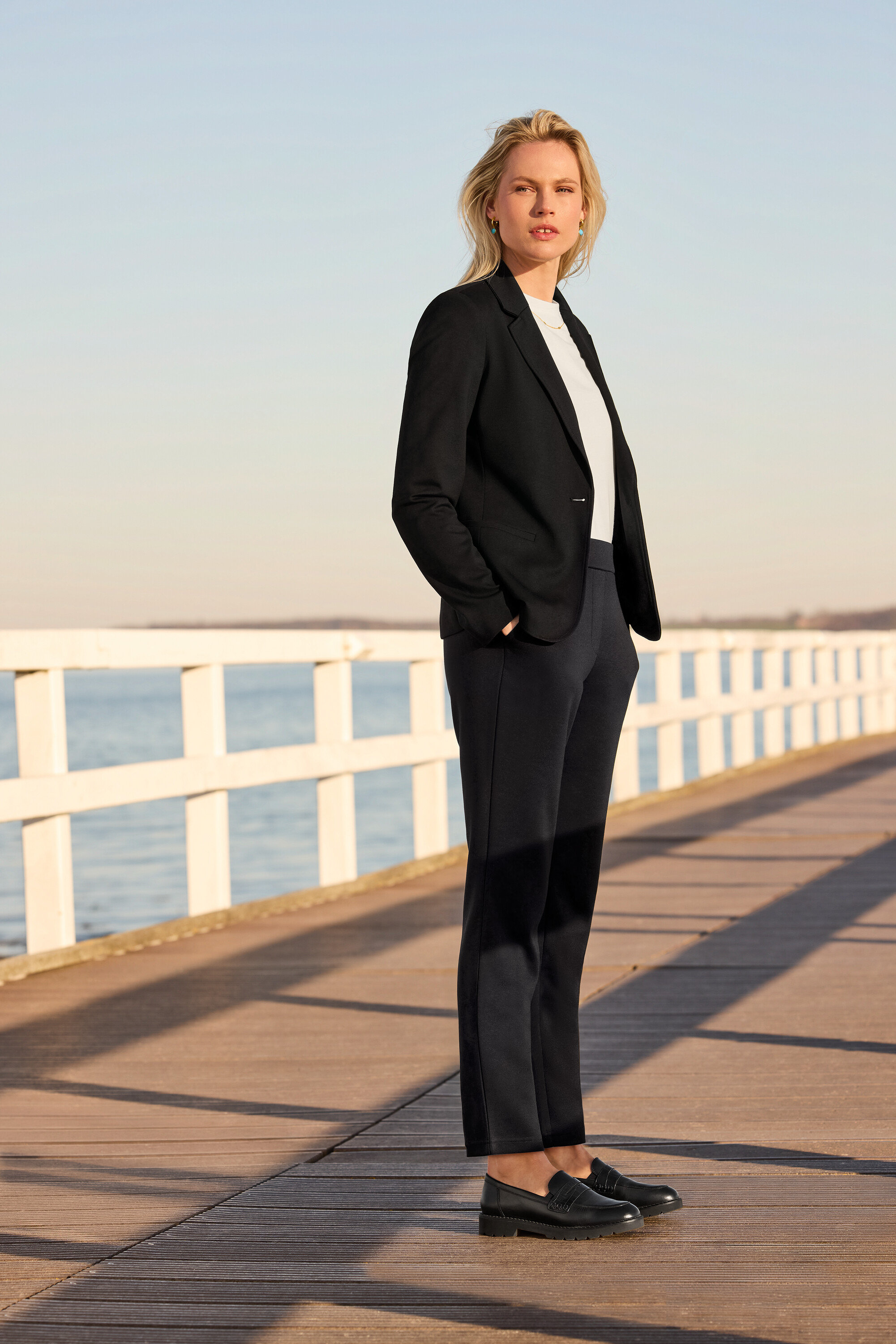 Eine Frau steht auf einem Pier und trägt eine Joggpants, ein Basic T-Shirt, weiß, einen Jerseyblazer und Leder-Loafer. Sie trägt Creolen mit Steinanhänger und eine Kette mit Navettes.