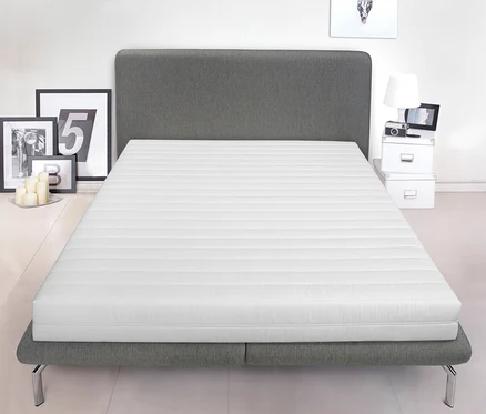Eine weiße Badenia-Kaltschaummatratze »Duo« liegt auf einem grauen Bett. Hinter dem Bett stehen Dekoartikel.