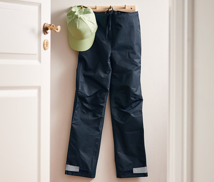 Eine Regenhose und eine grüne Kappe hängen an einer Garderobe. Auf der Fußmatte liegen eine Regenhose und Gummistiefel.