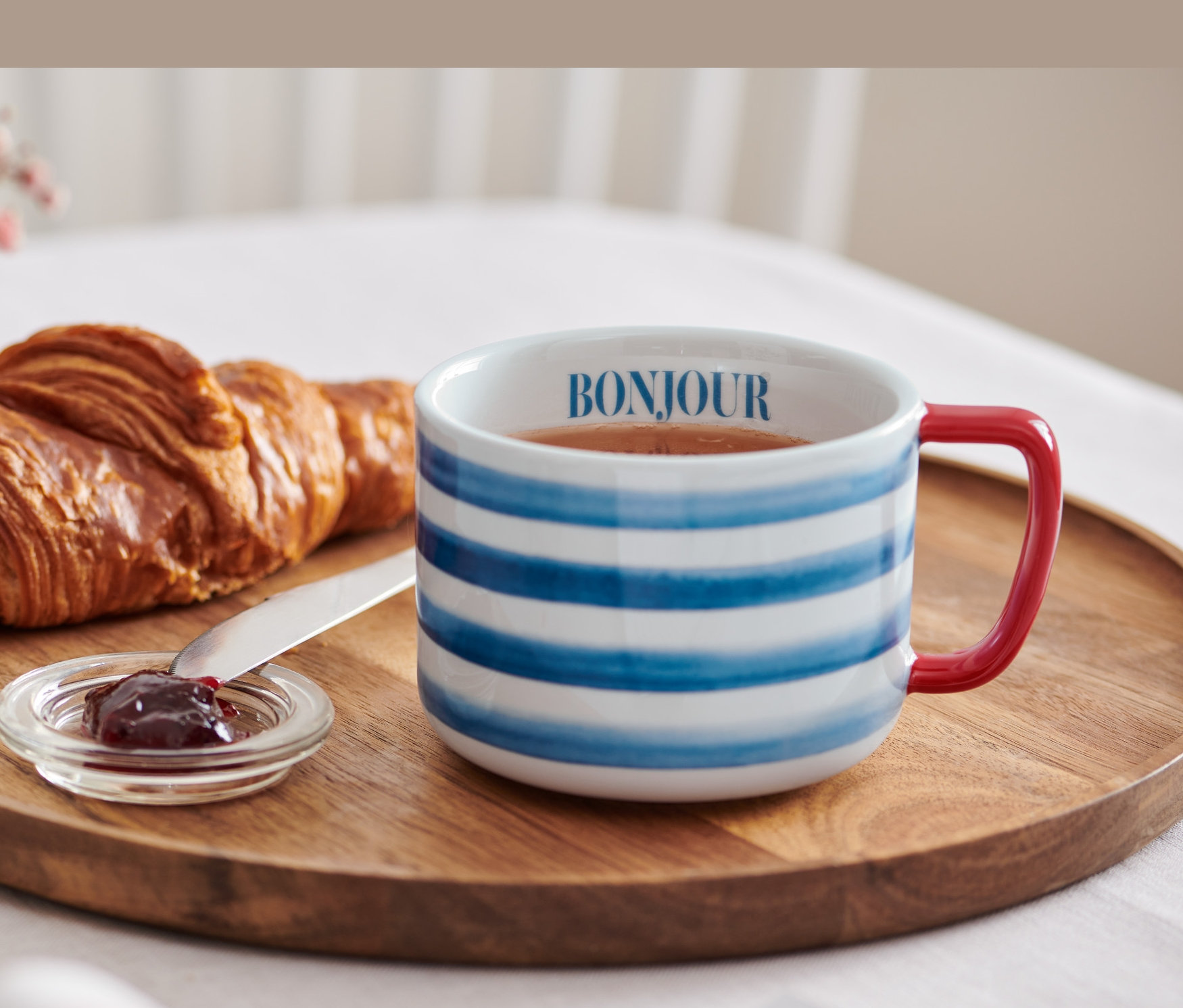 Auf einem Holztablett liegen ein Croissant, Marmelade und eine Jumbotasse mit der Aufschrift Bonjour.