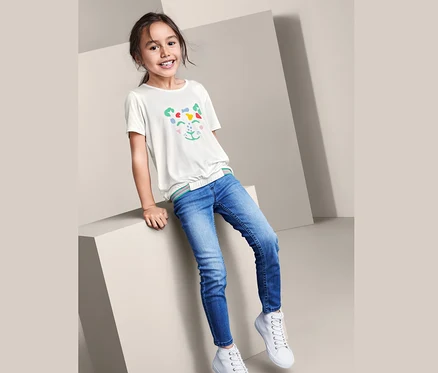 Ein Mädchen sitzt auf einem Würfel und trägt Jeans, weiße Turnschuhe und ein weißes T-Shirt in Schlupfform mit einem Tiergesicht.