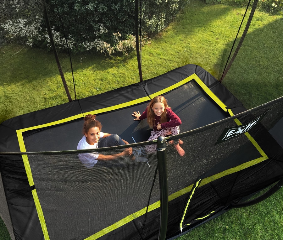 Zwei Mädchen sitzen in einem rechteckigen, schwarzen EXIT Silhouette Bodentrampolin mit gelber Umrandung im Rasen.