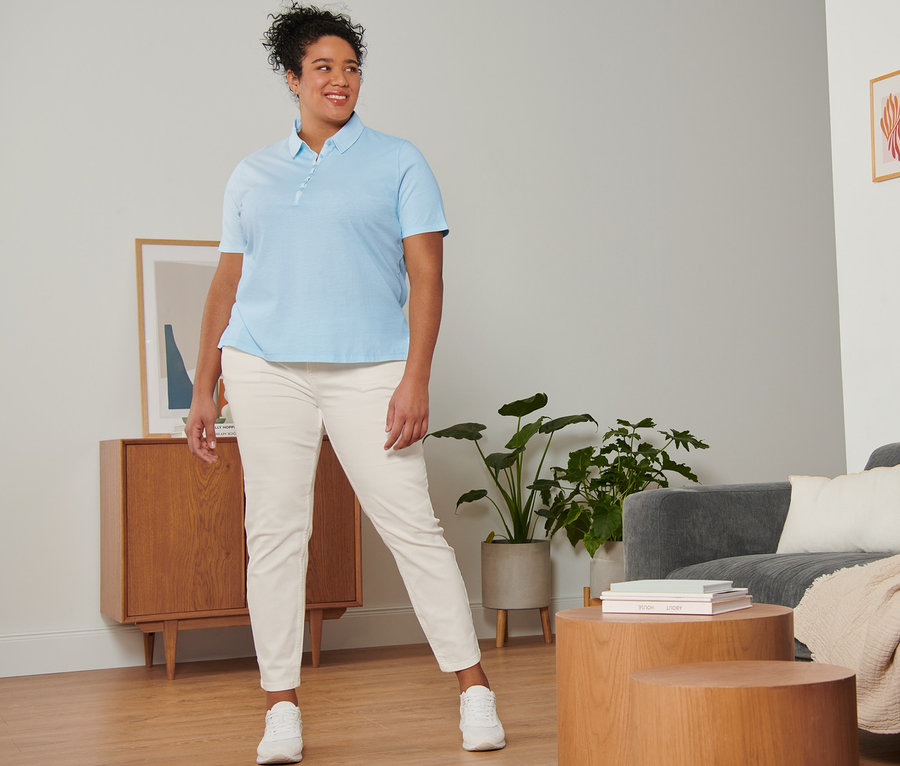 Frau posiert in cremeweißen Slimfit-Jeans, hellblauem Poloshirt und weißen Veloursleder-Sneakern.