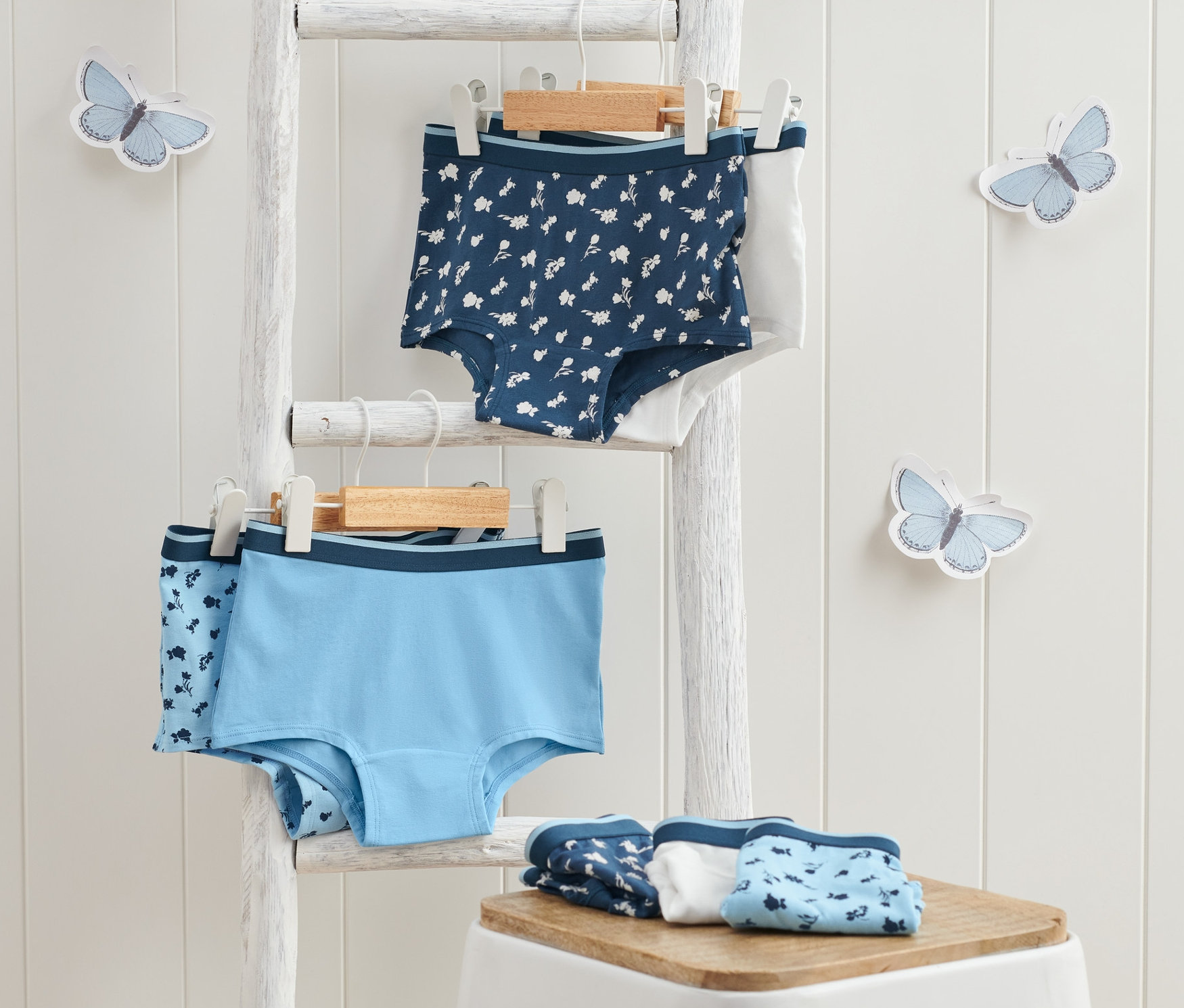 Zwei 7 Mädchen-Pantys, blau, hängen an einer weißen Leiter, weitere liegen gefaltet auf einem weißen Hocker.