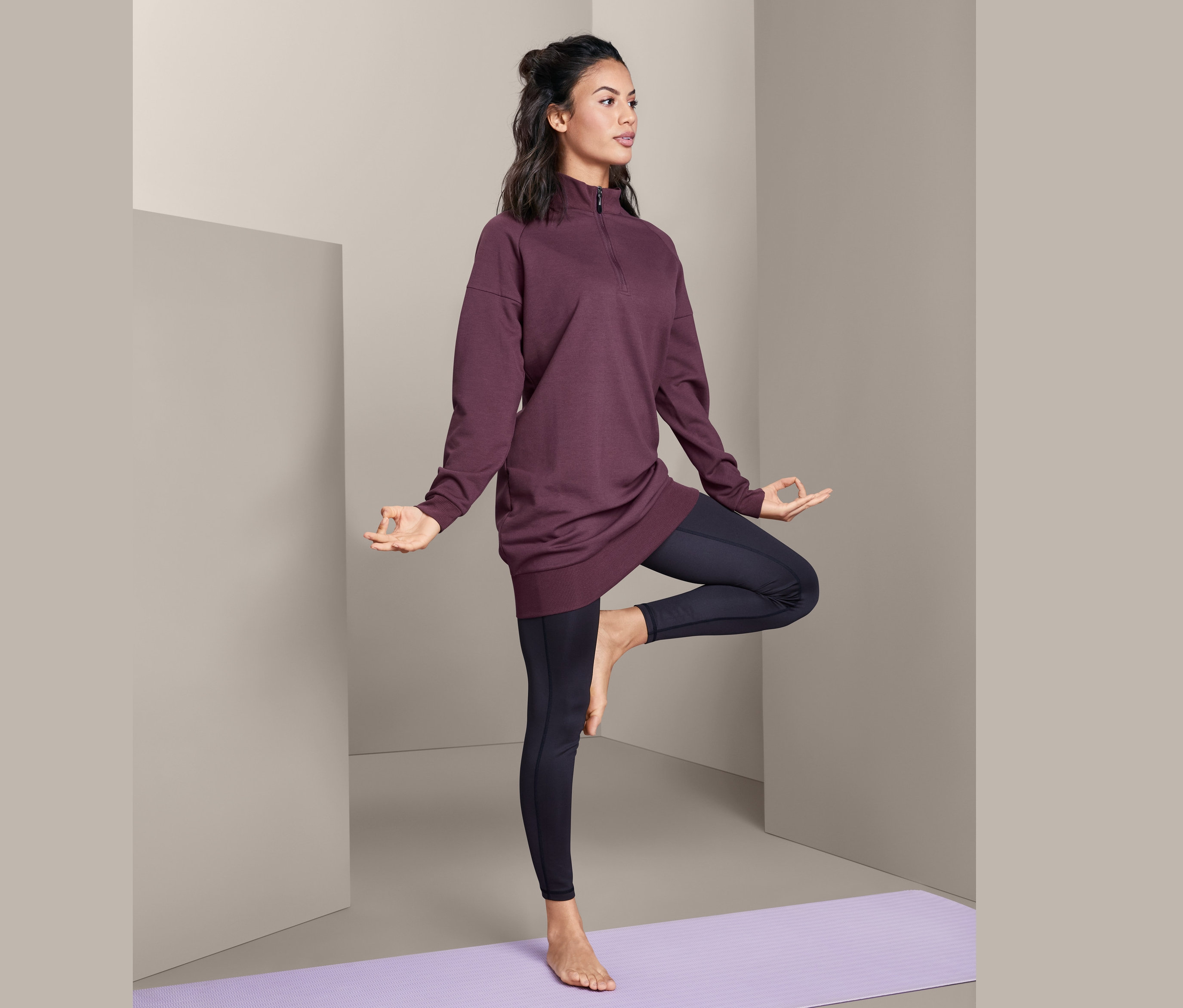 Frau im Yoga-Longshirt steht auf einer lila Yogamatte und macht die Baum-Pose.