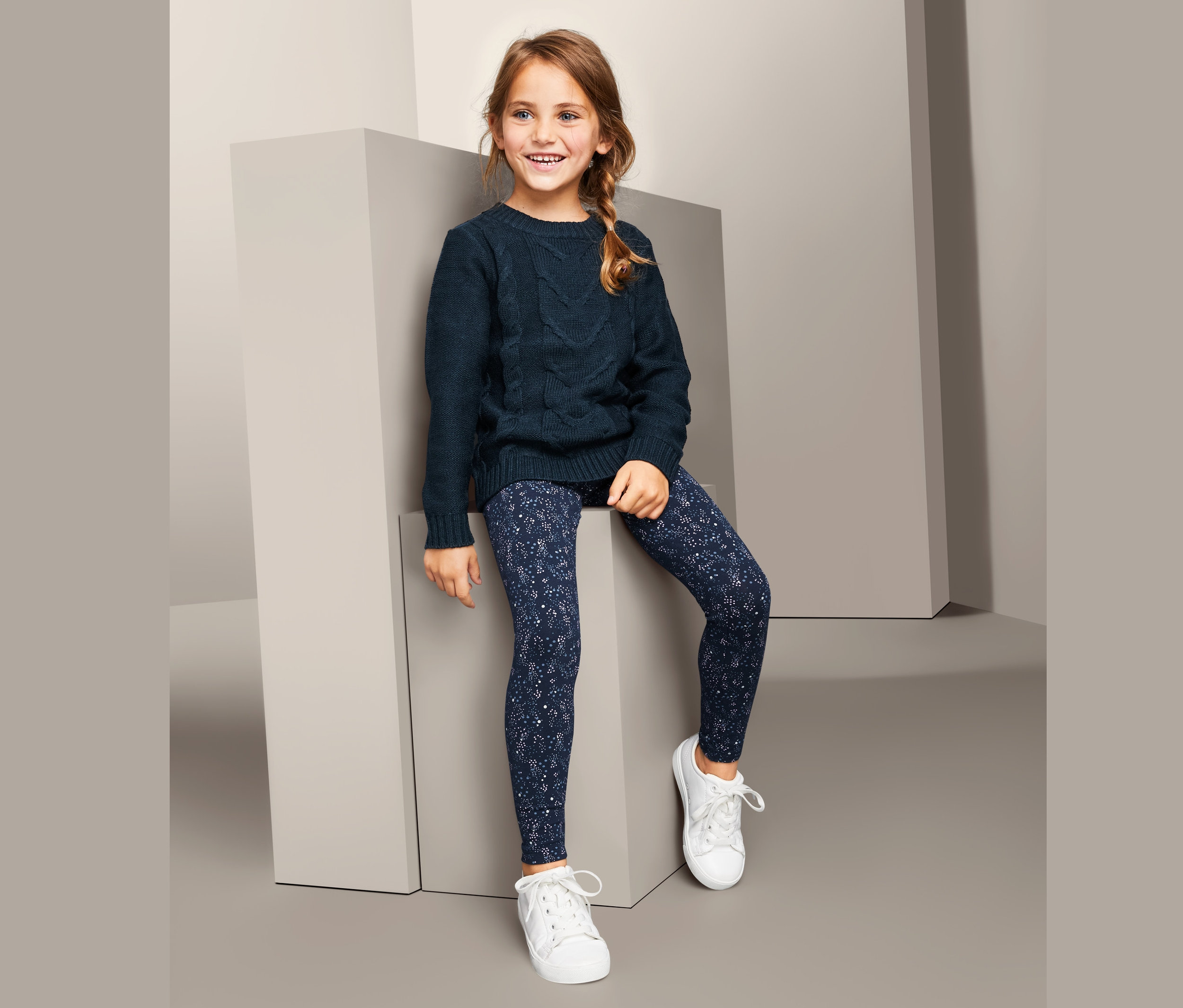 Lächelndes Mädchen sitzt auf einem grauen Würfel und trägt einen dunkelblauen Pullover und 3 Leggings.