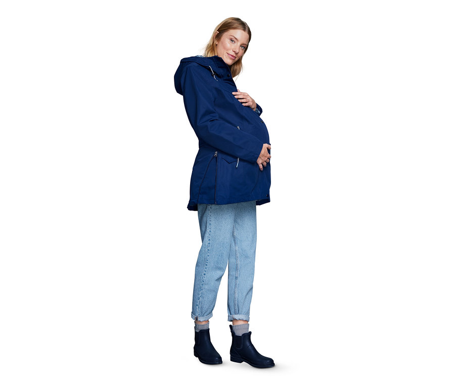 Schwangere Frau posiert in marineblauer Umstands-Regenjacke, blauen Jeans und marineblauen Stiefeln.