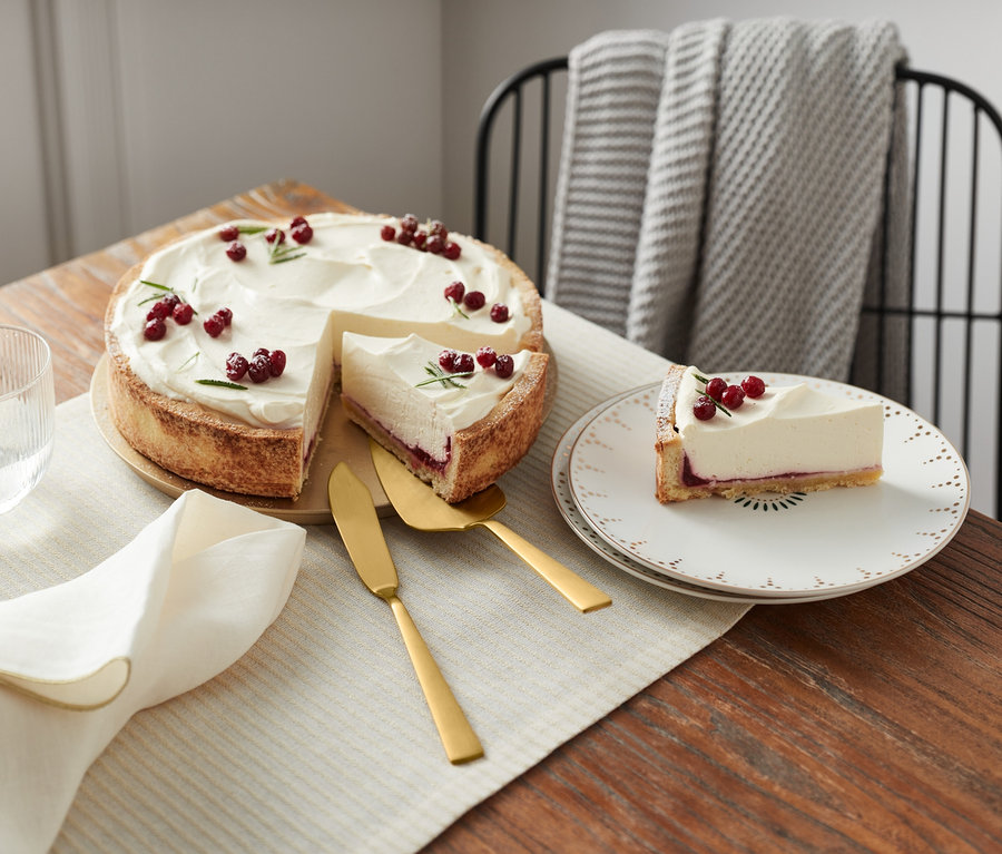 Torte mit weißem Guss und roten Früchten auf einem Holztisch. Ein 2-teiliges Tortenbesteck liegt neben der Torte.