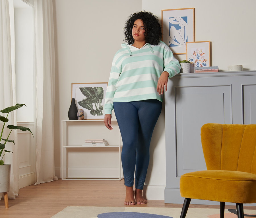 Eine Frau, die sich an eine Wand lehnt, trägt ein gestreiftes Kapuzen-Sweatshirt in Mint-Creme und Leggings in Jeansoptik.