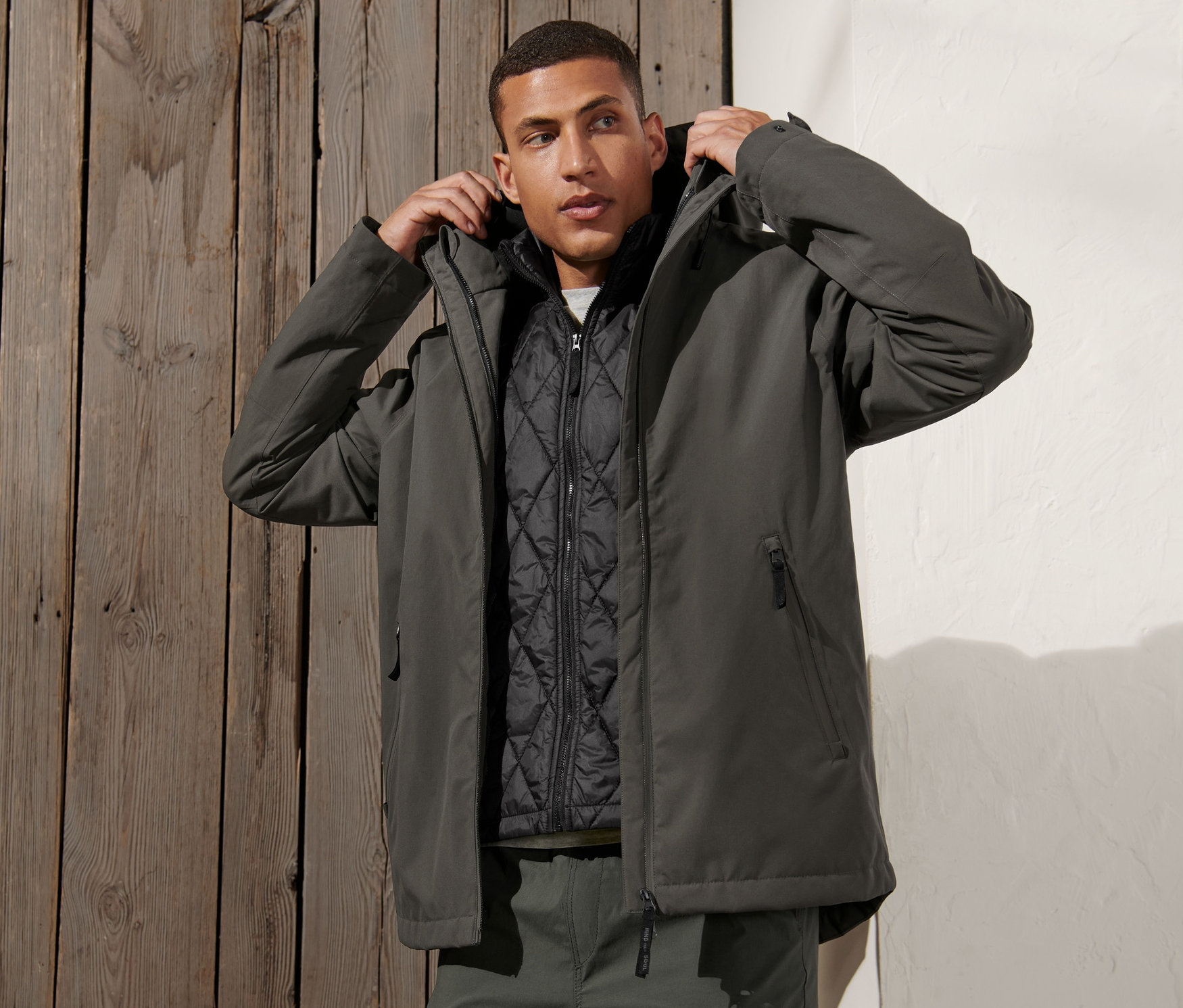 Mann justiert Kapuze der grüngrauen 3-in-1-Outdoorjacke mit Weste.