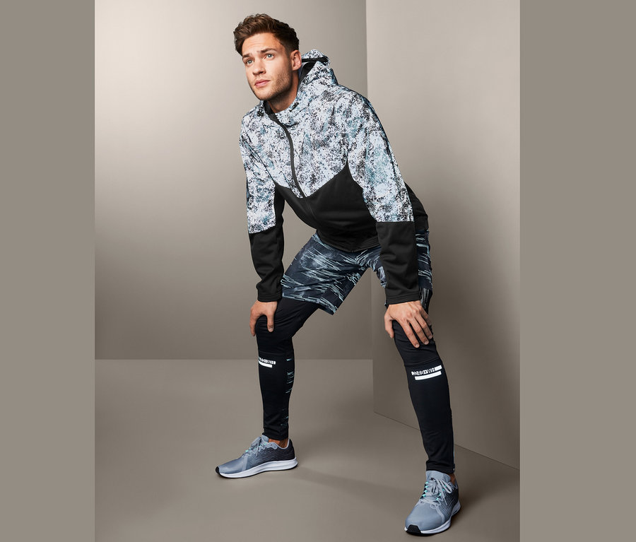 Mann mit aufgestützten Händen auf den Knien, trägt eine Softshelljacke mit dekorativen Reflektivelementen, Shorts und Leggings.
