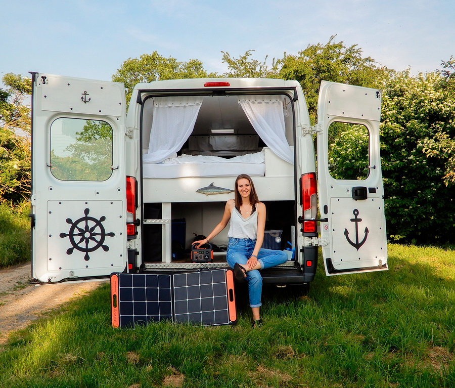 Junge Frau sitzt in offenem, weißen Wohnwagen auf Gras neben Solarpanel und Jackery Explorer 240.