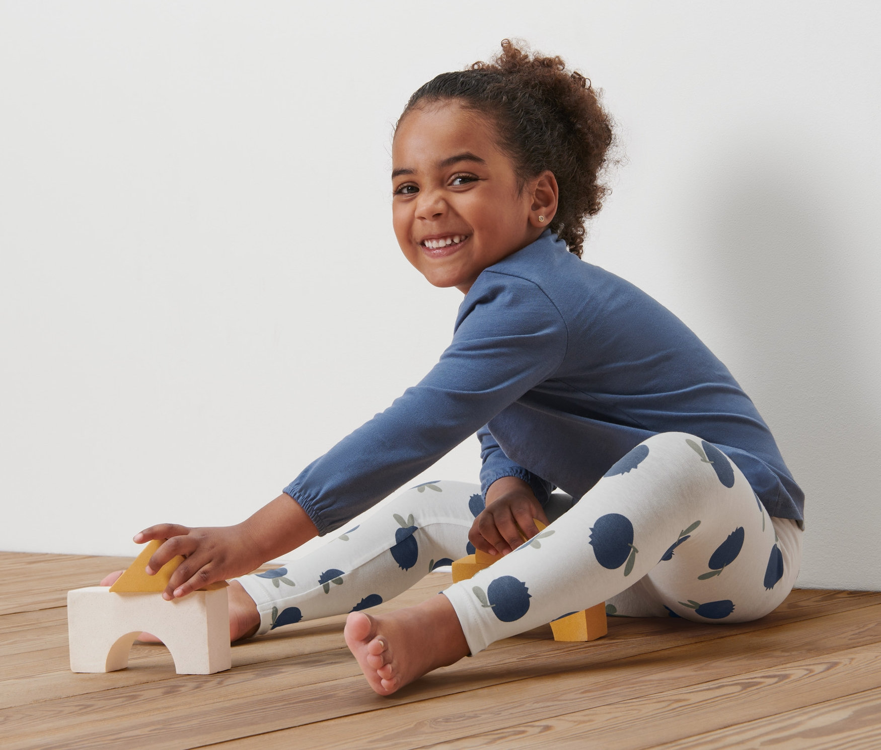 Ein lächelndes Mädchen sitzt auf einem Holzboden und spielt mit Holzspielzeug. Sie trägt ein blaues Oberteil und 5 Baby-und-Kleinkind-Leggings, Blaubeere.