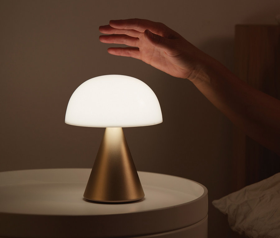Eine Hand greift nach der goldenen LEXON LED-Akku-Tischleuchte »Mina L«, die auf einem weißen Nachttisch neben einem Bett steht.